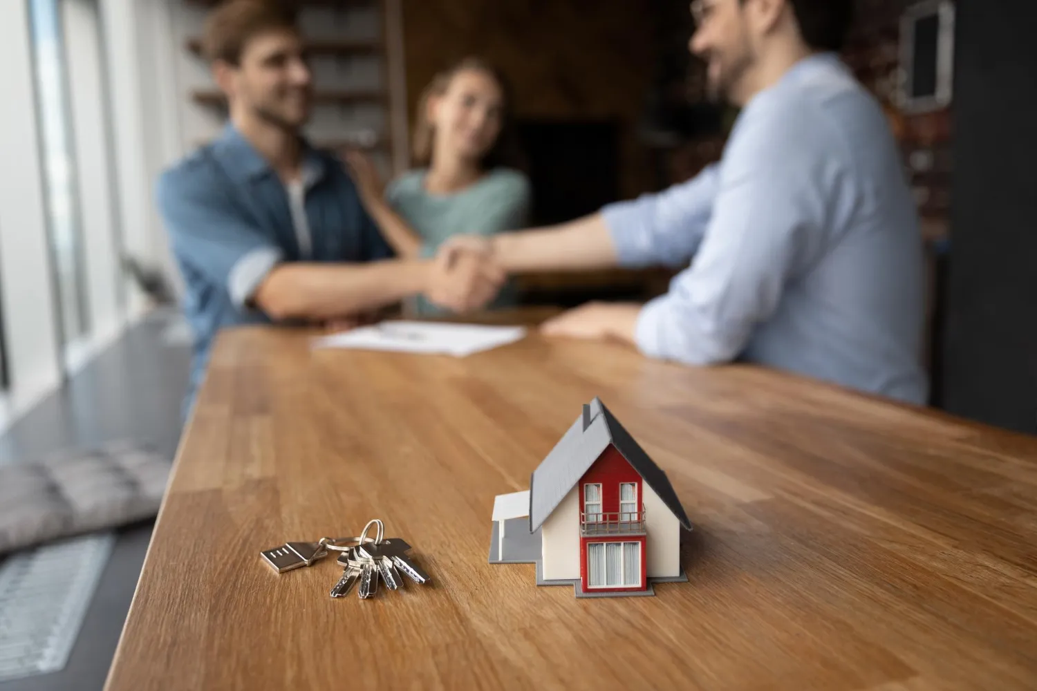 Gros plan sur une petite maison avec clés posées sur une table avec en arriere plan un couple vendant sa maison à une autre personne.
